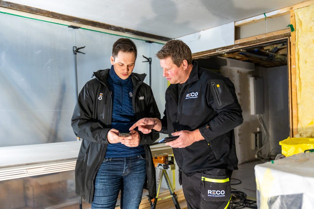To menn i arbeidsjakker fører prosjekt på en mobiltelefon i et delvis renovert rom, med byggeverktøy og materialer i bakgrunnen.