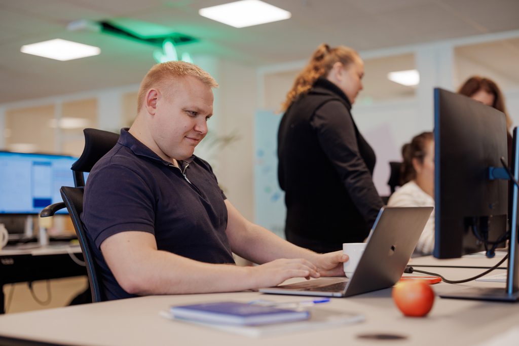 En mann i en blå poloskjorte er fokusert på den bærbare datamaskinen sin ved et skrivebord, omgitt av en dataskjerm, en bok og et eple. I bakgrunnen diskuterer to kvinner implikasjonene av «skatt på bonus» med animerte bevegelser.