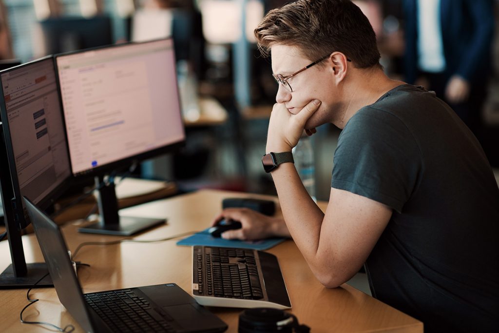 En person med briller jobber intenst ved et skrivebord med to skjermer og en bærbar datamaskin, og analyserer omhyggelig data knyttet til finansiell kriminalitet i kontormiljøer.