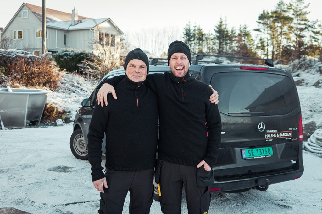 To smilende menn i svarte gensere står arm i arm i en snødekt hage foran en svart varebil. Klare for å føre prosjekt i Tripletex