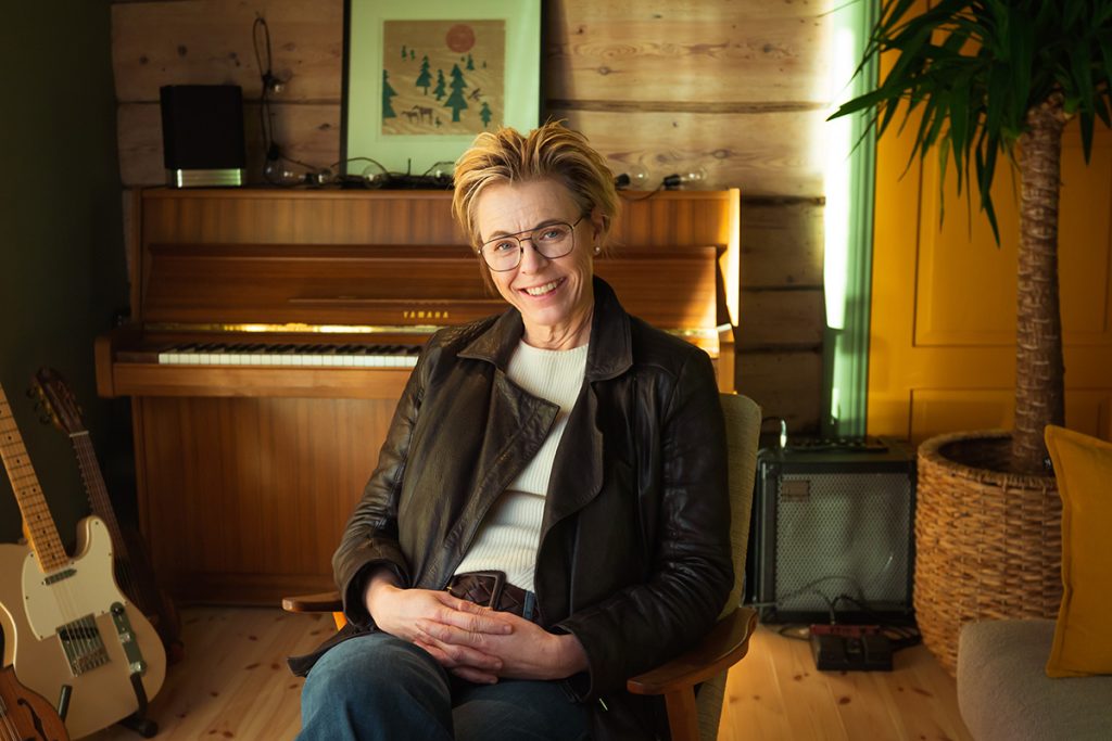 Person med kort hår og briller som sitter i et rom med piano, gitar og forsterker. Hun har på seg en skinnjakke og smiler til kameraet. Et innrammet kunstverk er på veggen bak.
