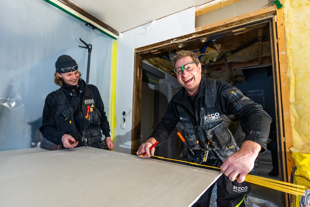 To bygningsarbeidere fra RECO måler og skjærer et gipspanel, begge iført svarte arbeidsuniformer og vernebriller, inne i et delvis renovert rom.