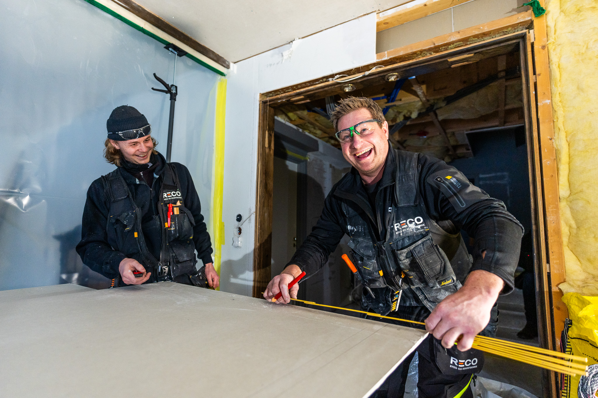 To bygningsarbeidere fra RECO måler og skjærer et gipspanel, begge iført svarte arbeidsuniformer og vernebriller, inne i et delvis renovert rom.