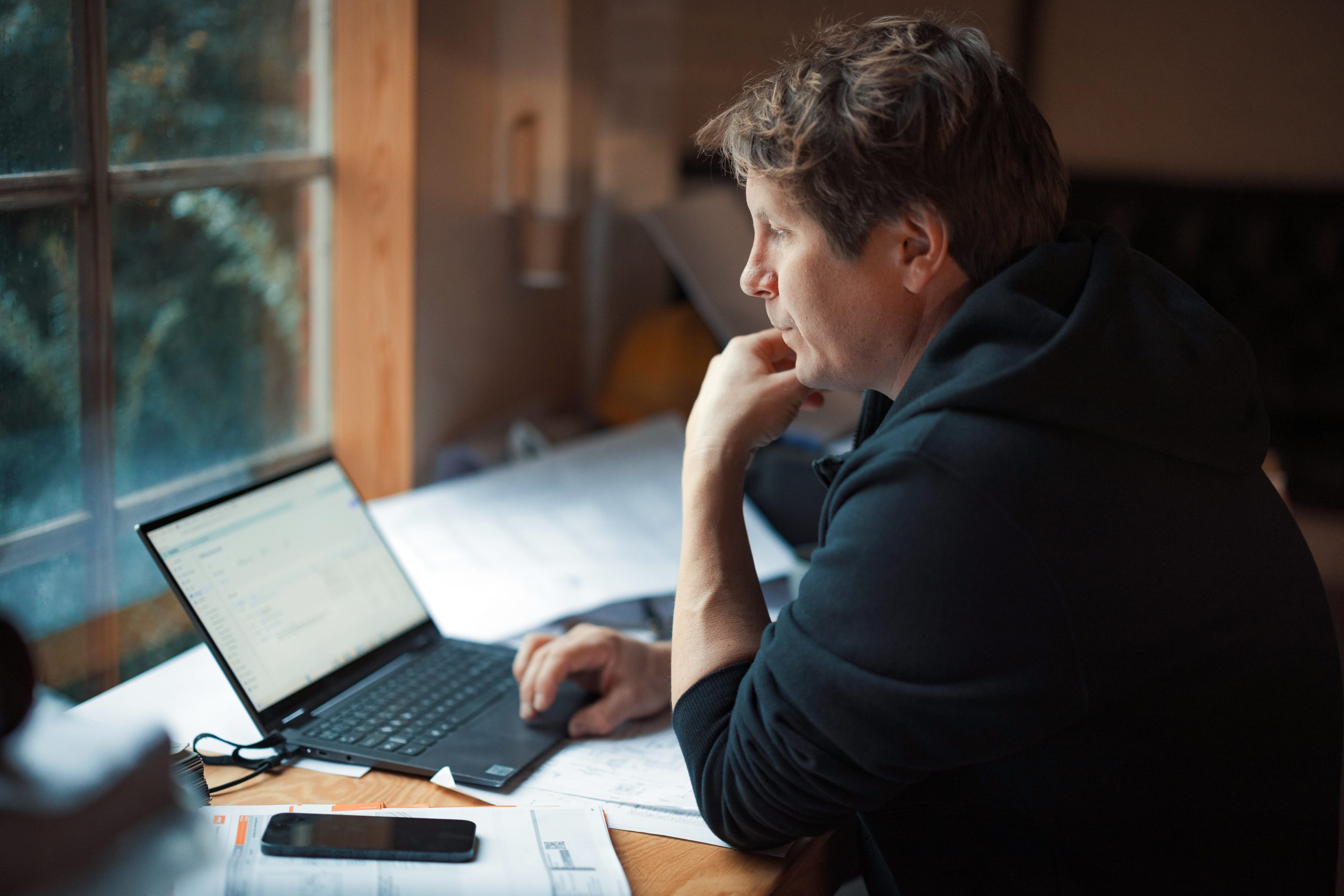 Mann i svart hettegenser sitter ved et skrivebord og fører klimaregnskap på en bærbar skjerm med en smarttelefon og papirer i nærheten.