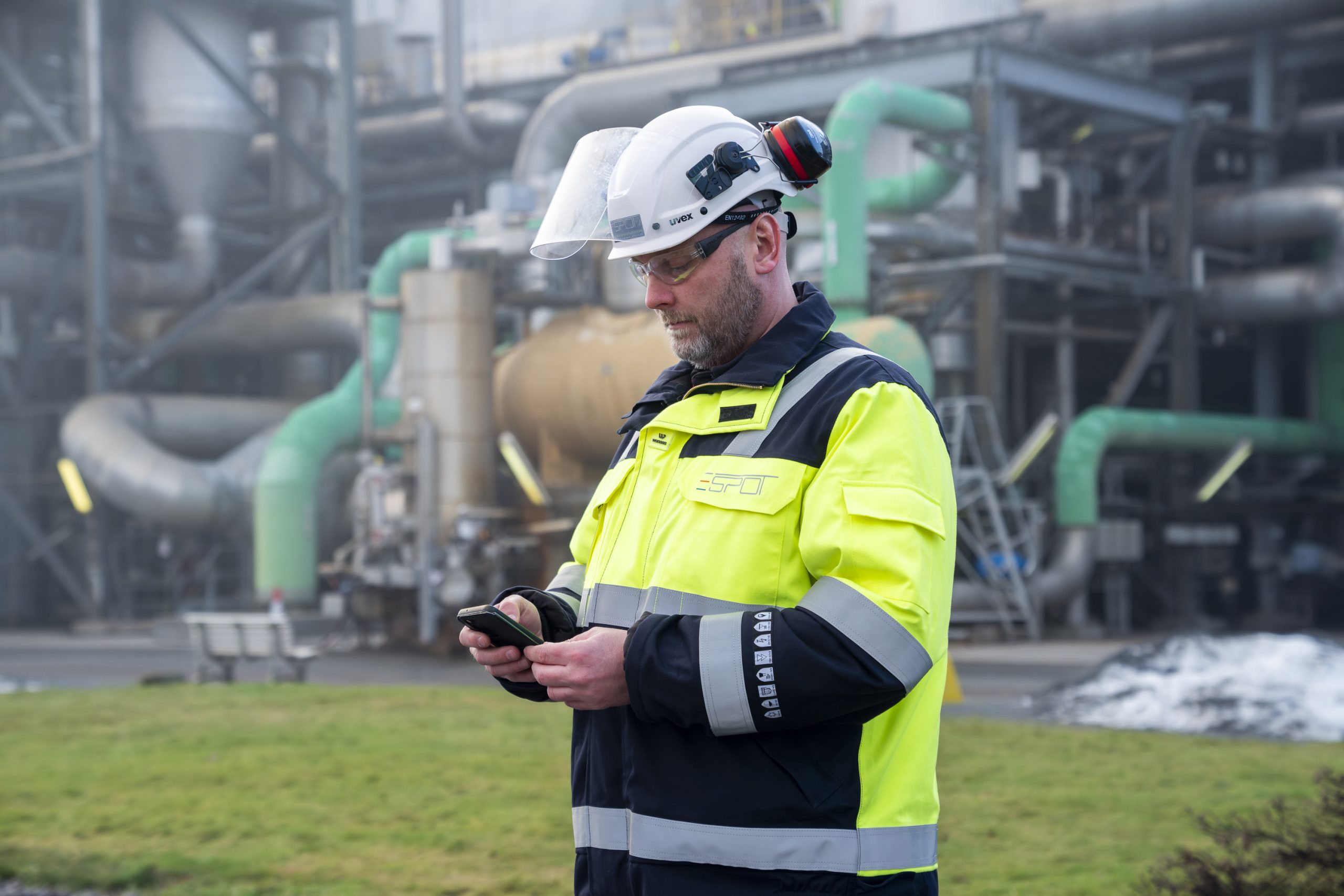 En person i gul og blå sikkerhetsjakke med hjelm og briller bruker Tripletex på mobil. Industrielt utstyr og rør er synlig i bakgrunnen.