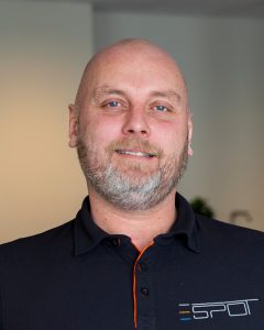 Mann med skjegg iført svart poloskjorte står innendørs og smiler lett. Jon-Henrik i Spot AS.