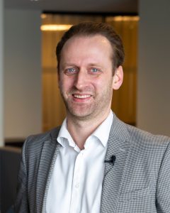 En person i grå dress og hvit skjorte smiler. Bård i OneAccounting står innendørs, uskarp bakgrunn.