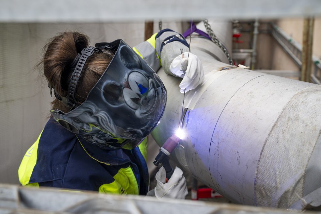 En person som har på seg verneutstyr sveiser et stort metallrør ved hjelp av en sveisebrenner og vinkelhodespeil.