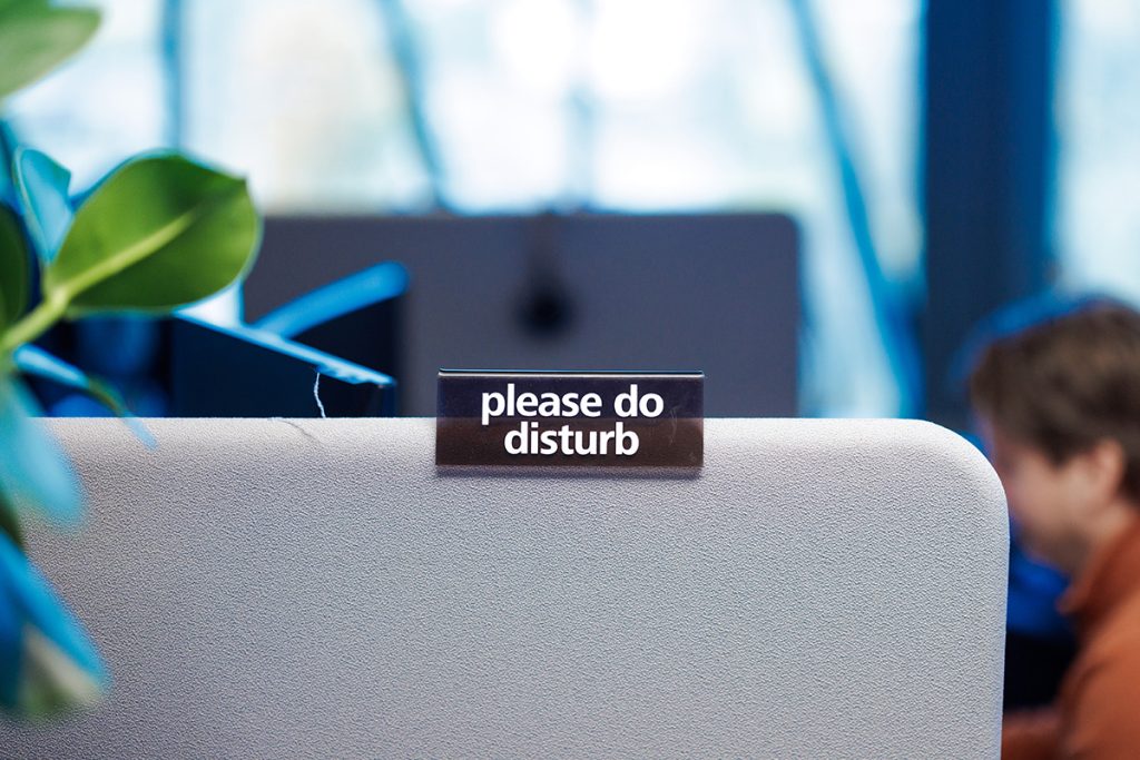 Et skilt med teksten "please do disturb" er festet til toppen av en grå kontorpartisjon, med en person som jobber i uskarp bakgrunn.