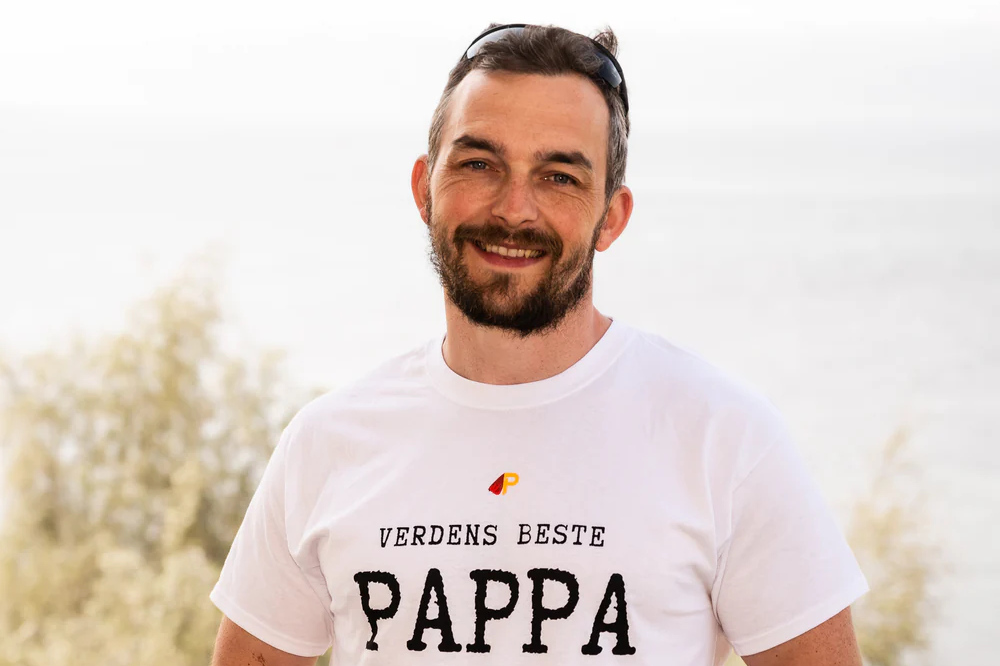 En mann med skjegg og solbriller på hodet smiler mot kameraet, iført en hvit t-skjorte med påskriften "Verdens Beste Pappa".