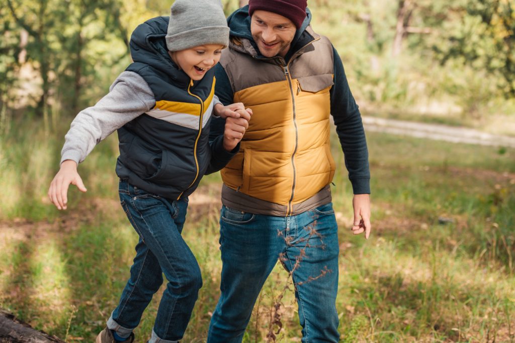 Et barn og en voksen i jakker går hånd i hånd gjennom en skog, og barnet ser ut til å være midt i trinnet på en tømmerstokk.