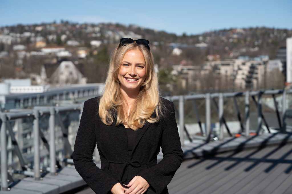 En kvinne med langt blondt hår iført en svart frakk står på et tak og drømmer om feriepenger, med et bybilde og åser i bakgrunnen.
