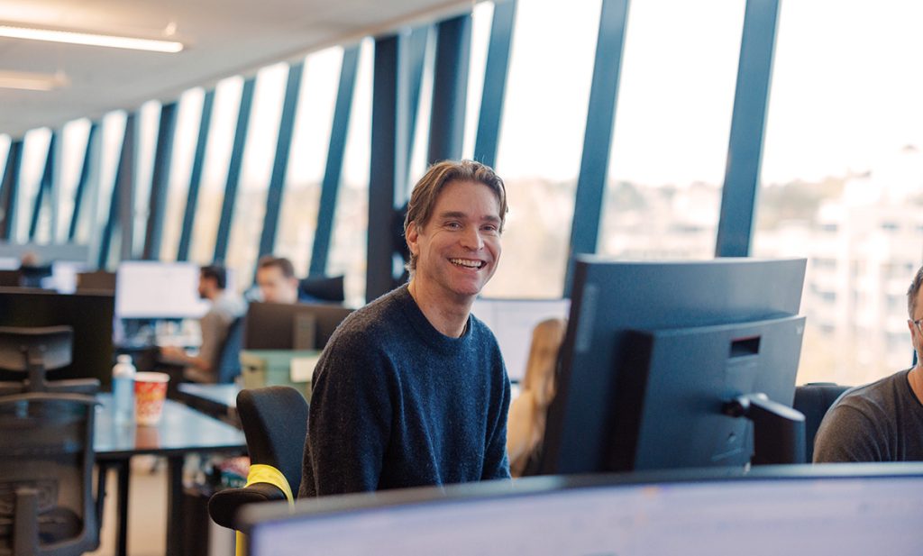 En person sitter ved et skrivebord på et åpent kontor med flere skjermer og smiler til kameraet. Flere kolleger jobber i bakgrunnen med store vinduer langs veggen.