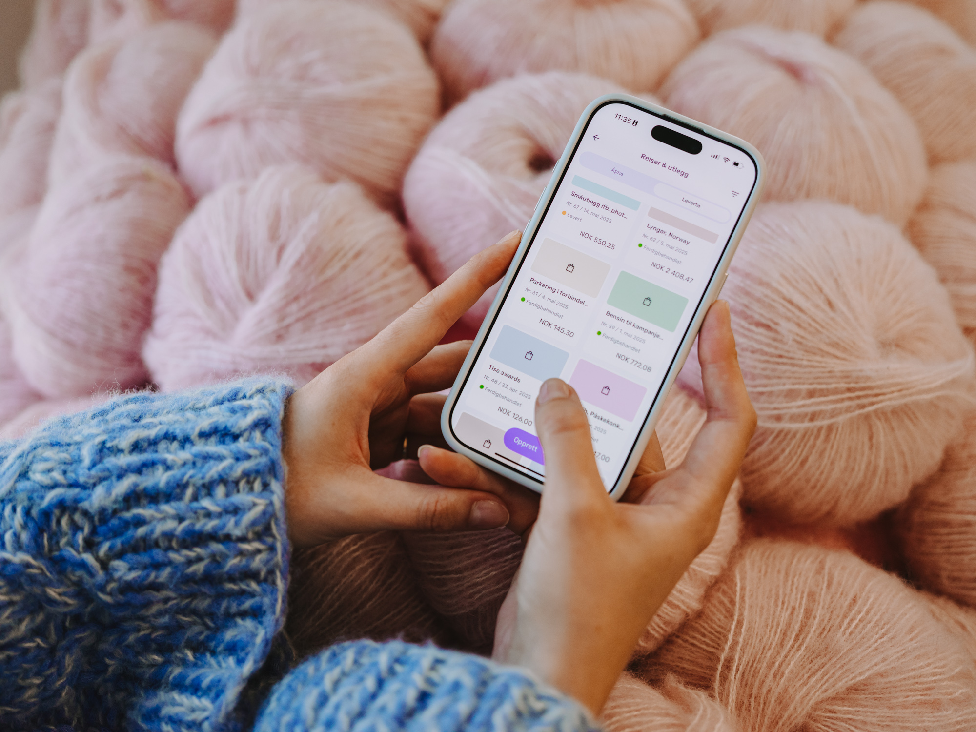 Hender holder en smarttelefon som viser Tripletex-appen, med rosa garnnøster i bakgrunnen.