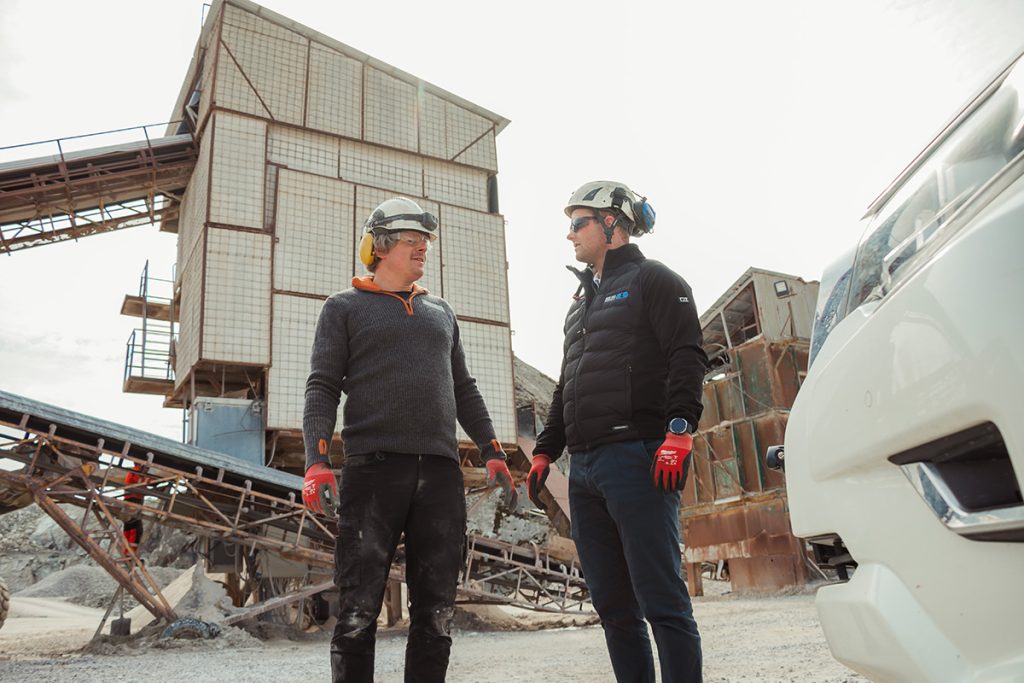 To menn med vernehjelmer og verneutstyr står og snakker på et industriområde med metallkonstruksjoner og utstyr i bakgrunnen.