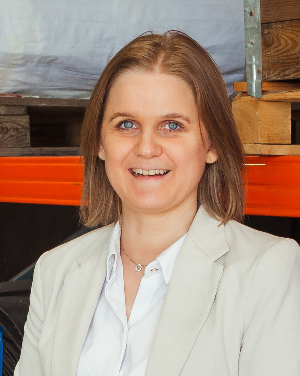 En kvinne med rett lysebrunt hår, iført en lys blazer og hvit skjorte, smiler i et industri- eller lagermiljø.