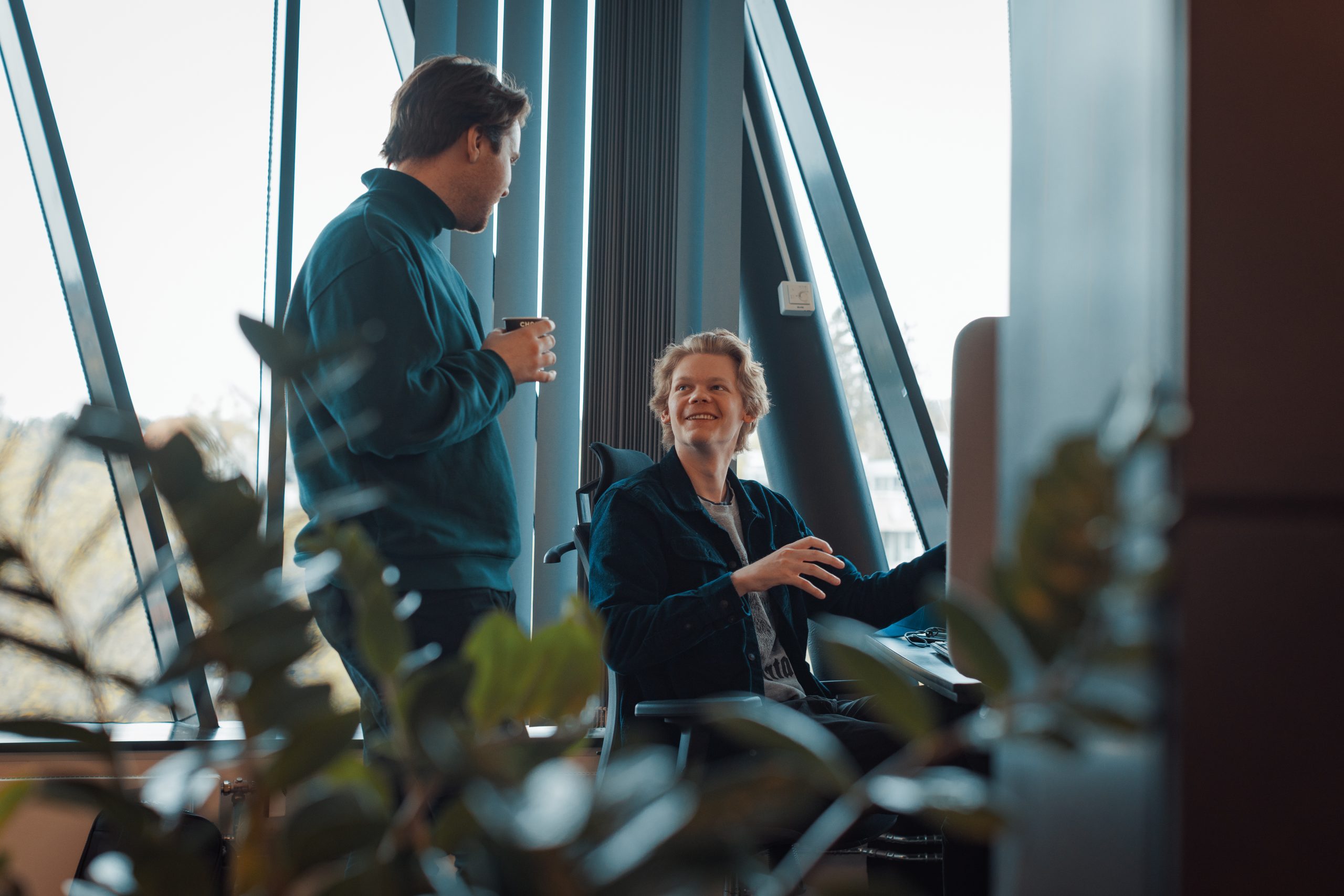 To personer fører en samtale på et kontor. Den ene sitter ved et skrivebord med en datamaskin, mens den andre står ved siden av og holder en kopp.