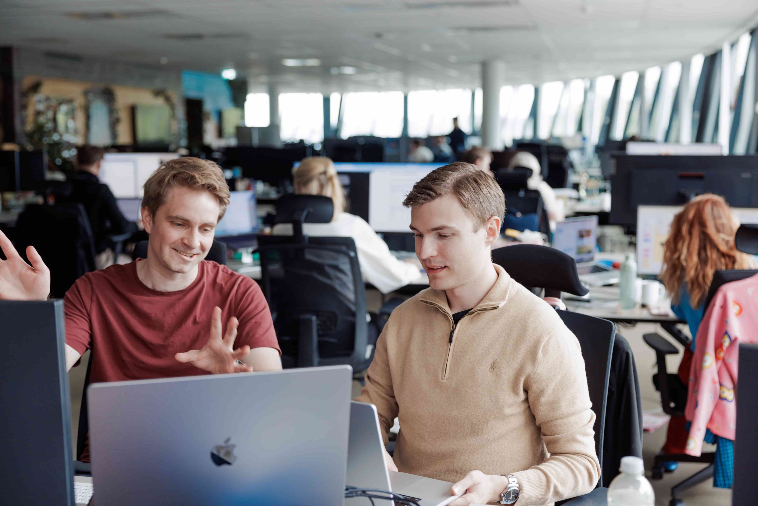 To menn sitter ved et skrivebord på et moderne kontor og diskuterer mens de ser på en bærbar datamaskin. Andre ansatte arbeider ved datastasjoner i bakgrunnen.