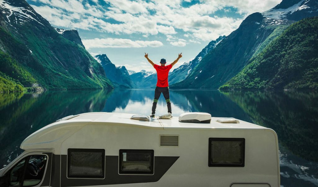 En person står på toppen av en bobil foran en fjord, til artikkel om huskeliste før man logger av i ferien.