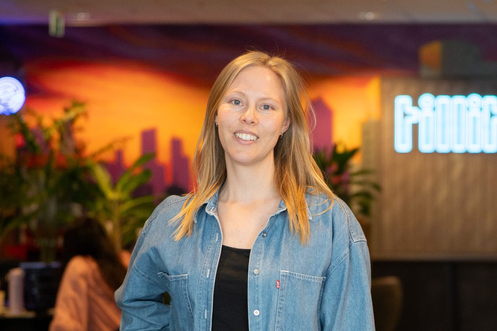 En kvinne med blondt hår i dongeriskjorte står innendørs og smiler til kameraet. Det er planter og uskarpe mennesker i bakgrunnen med fargerik veggkunst.
