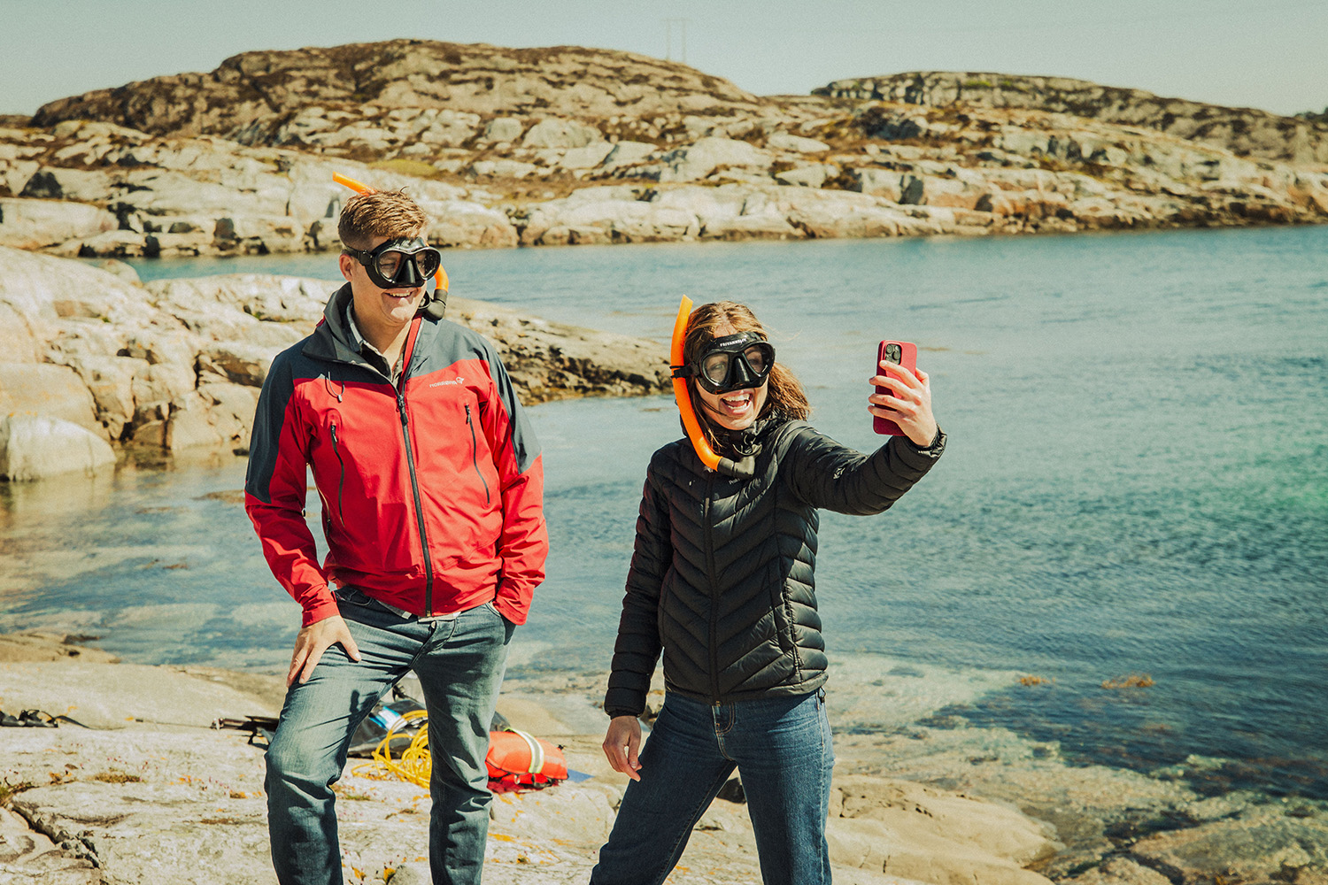 To personer iført snorkelmasker og jakker står på svaberg ved vannet, mens den ene tar en selfie med smarttelefon.