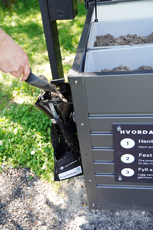 En person fyller en svart plastpose med sand fra en metalldispenser ved hjelp av en liten håndspade utendørs.