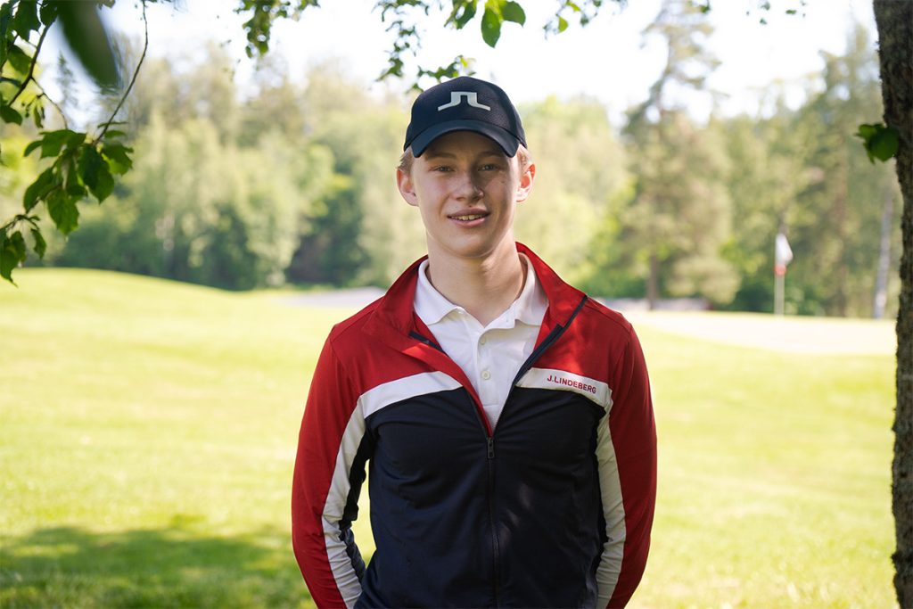 En ung mann med svart lue og rød og svart jakke står utendørs en solskinnsdag, med en golfbane og trær i bakgrunnen.
