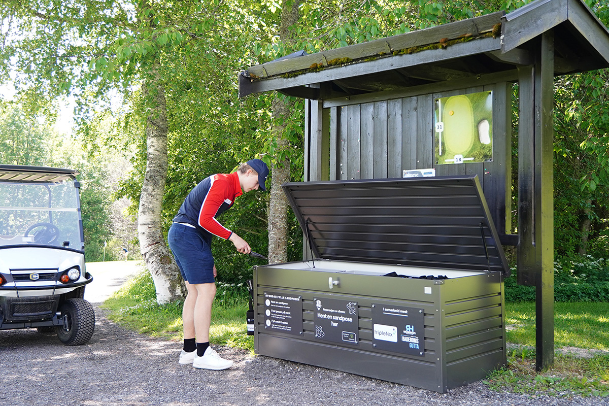 En person står ved siden av en åpen oppbevaringsboks for utstyr under et tak i nærheten av en golfbil på en sti med trær.