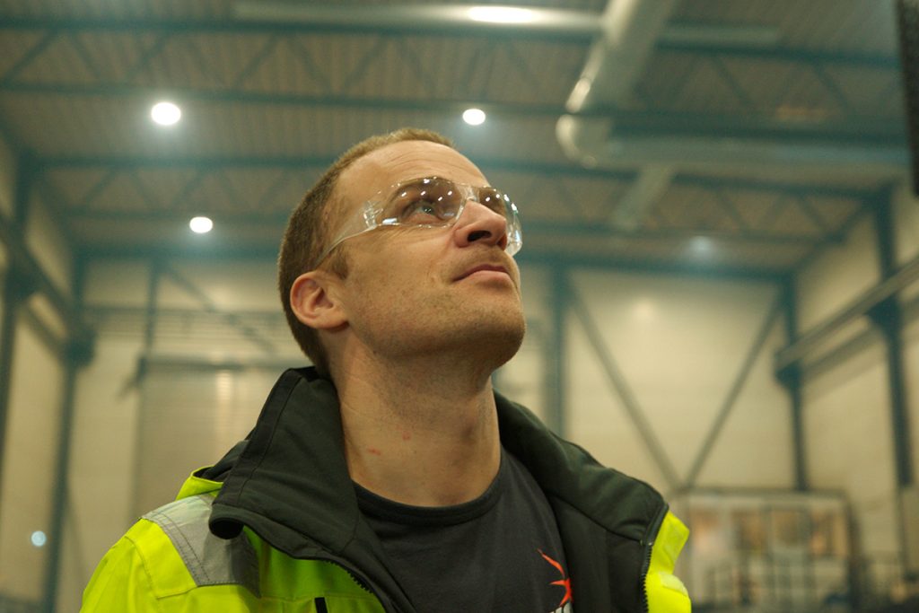 En mann iført vernebriller og refleksvest ser opp i en industribygning.