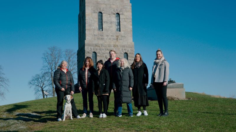 En kvinne med rett lysebrunt hår, iført en lys blazer og hvit skjorte, smiler i et industri- eller lagermiljø.