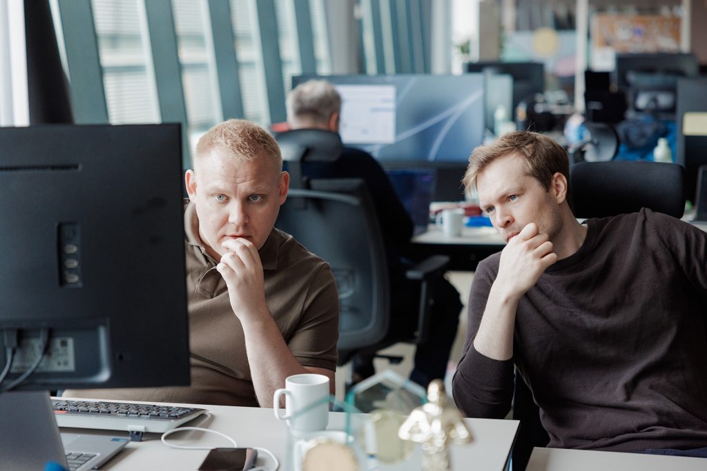 To menn sitter ved et skrivebord på et kontor og ser konsentrert på en dataskjerm mens de prøver å forstå regnskapstall. En annen person arbeider ved en datamaskin i bakgrunnen.