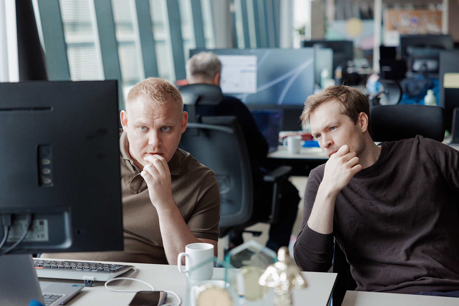 To menn sitter ved et skrivebord på et kontor og ser konsentrert på en dataskjerm mens de prøver å forstå regnskapstall. En annen person arbeider ved en datamaskin i bakgrunnen.