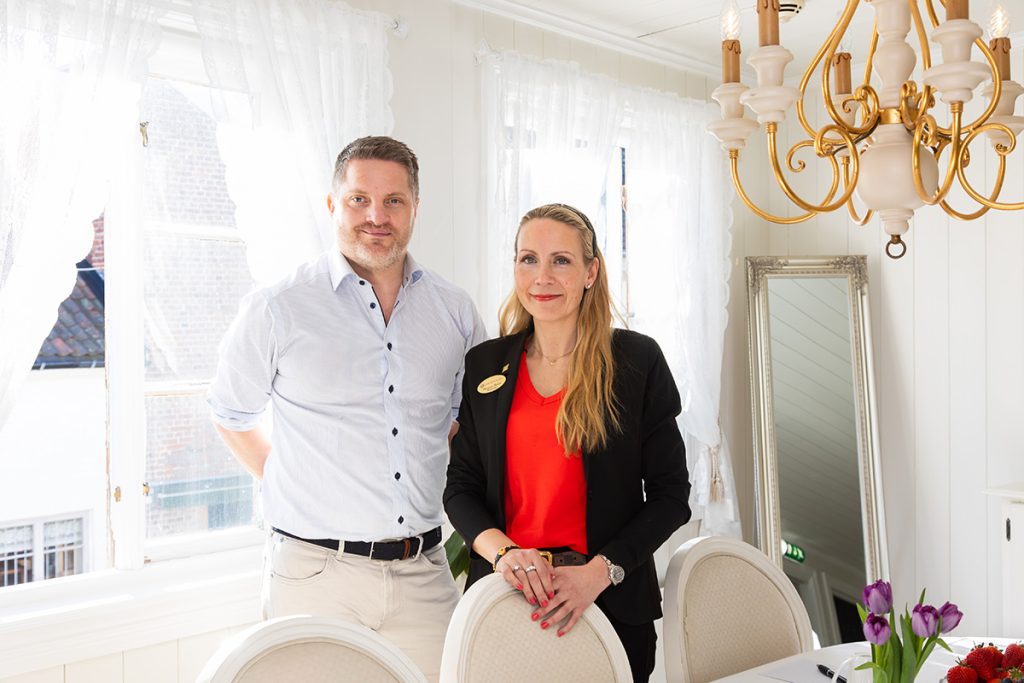 En mann og en kvinne står side om side i en lys, elegant spisestue med hvit innredning, lysekrone og en vase med lilla tulipaner på bordet.