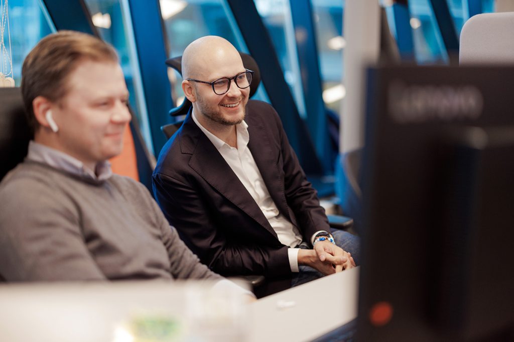 To menn sitter side om side ved et skrivebord og ser på en dataskjerm. Den ene har genser og AirPods, den andre har briller og blazer.
