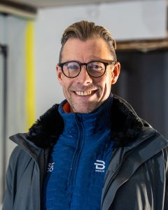 En mann med briller og bakoverslått hår smiler, iført blå jakke med høy krage og grå frakk, og står innendørs med uskarp bakgrunn.