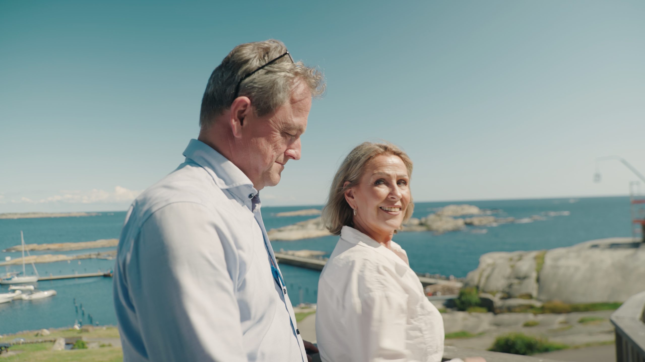 To voksne mennesker går utendørs nær kysten en solskinnsdag, med båter, svaberg og vann i bakgrunnen - bare en kort spasertur fra Gamlebyen Hotell.