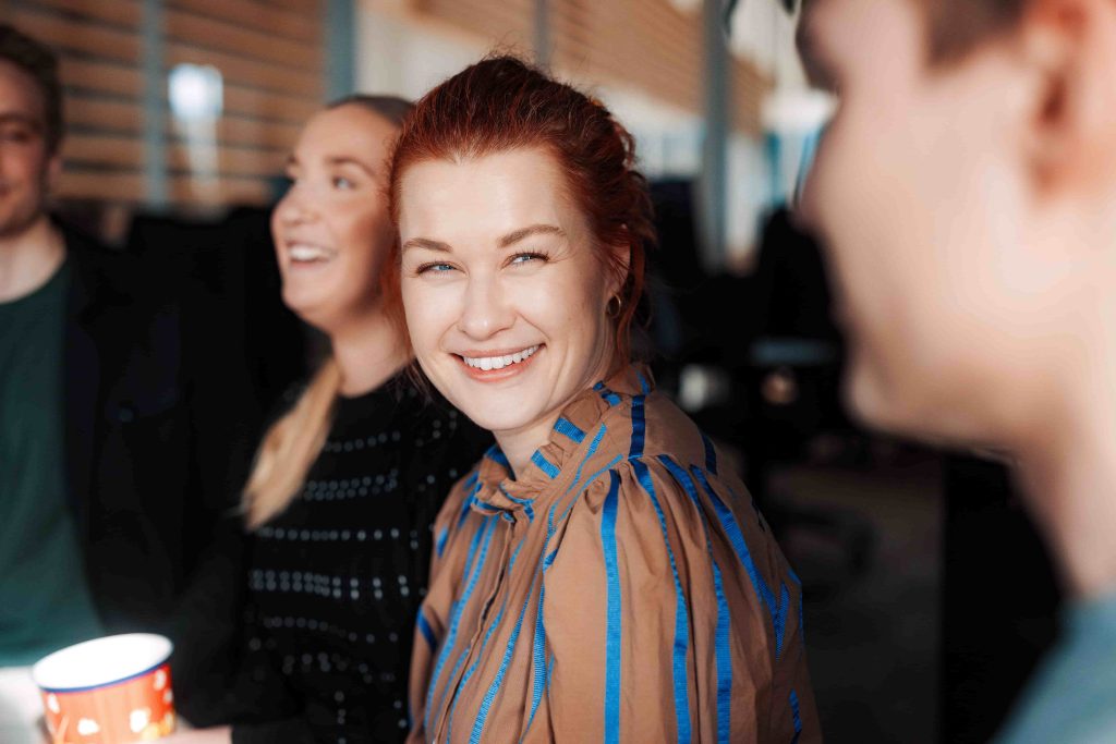 En kvinne med rødt hår smiler til kameraet mens hun sitter sammen med tre andre personer i et uformelt innemiljø.