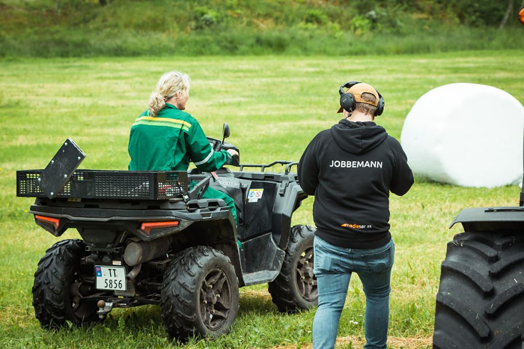 En grønnkledd person kjører ATV på gress, mens en annen person står i nærheten iført hodetelefoner og en svart jakke merket "JOBBEMANN". Et stort hvitt objekt er i bakgrunnen.
