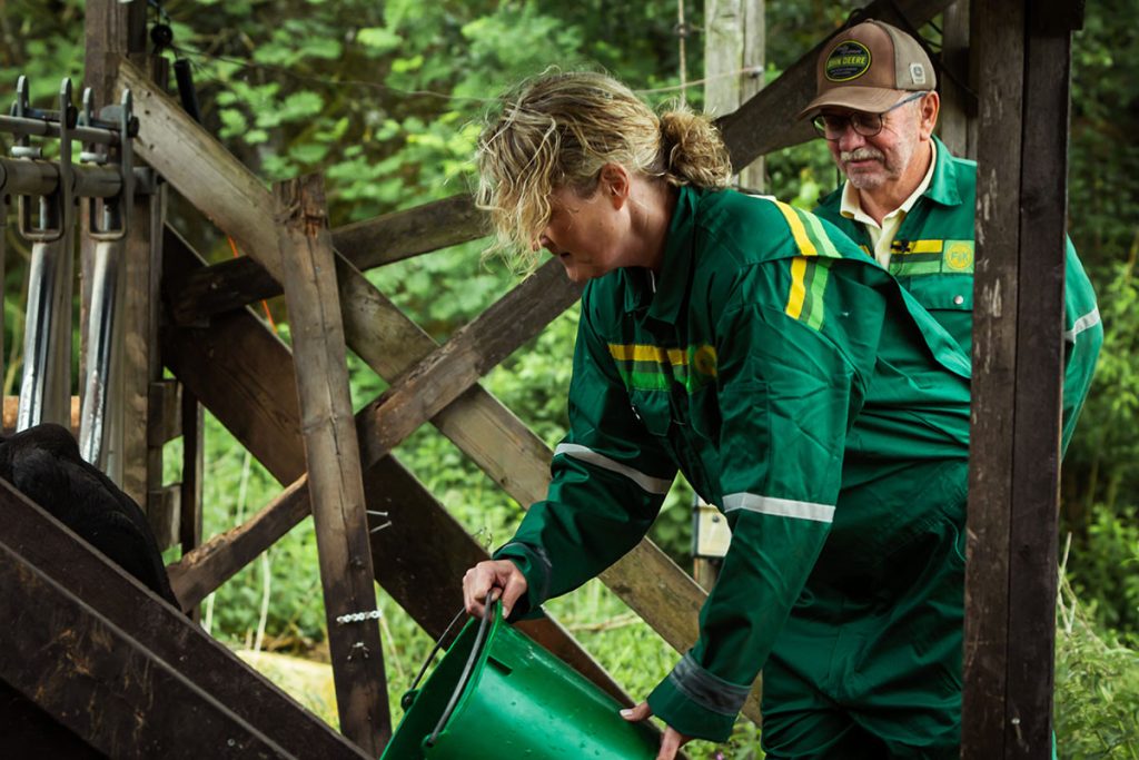 To personer i grønne kjeledresser arbeider på en gård; den ene skjenker fôr fra en grønn bøtte mens den andre observerer. I bakgrunnen skimtes trebjelker og grønne planter.
