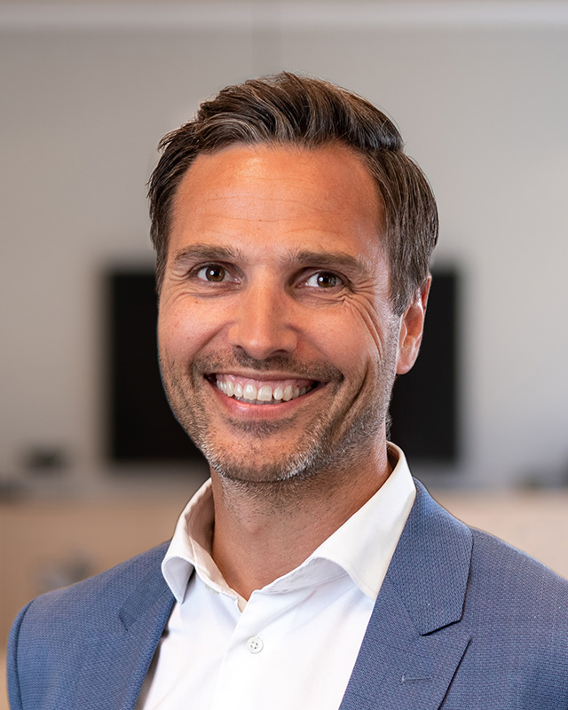 En mann i blå dressjakke og hvit skjorte smiler til kameraet i et kontormiljø med uskarp bakgrunn.