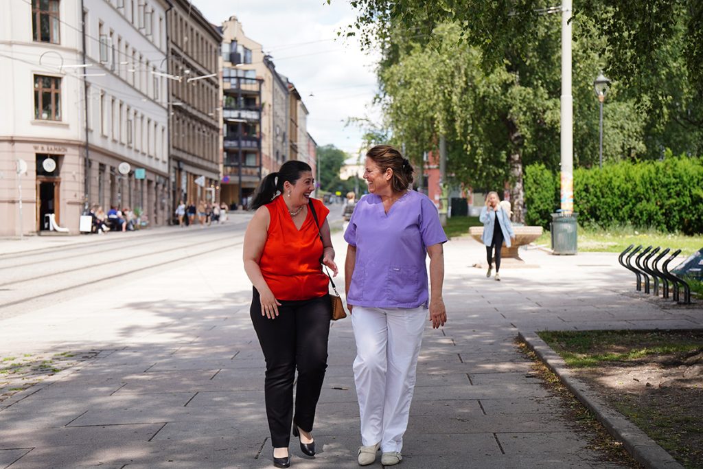 To kvinner, den ene i rød topp og den andre i lilla helseklær, går og snakker sammen på et solfylt fortau i en by med bygninger, benker og trær i bakgrunnen. Montebello Consulting og Grunerløkka Tannhelsesenter