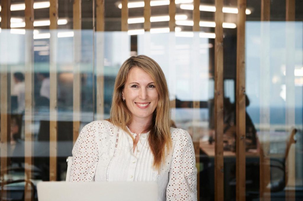 En kvinne med langt, blondt hår sitter smilende ved et skrivebord med en bærbar datamaskin i et moderne kontor med glassvegger og vertikale trelister.