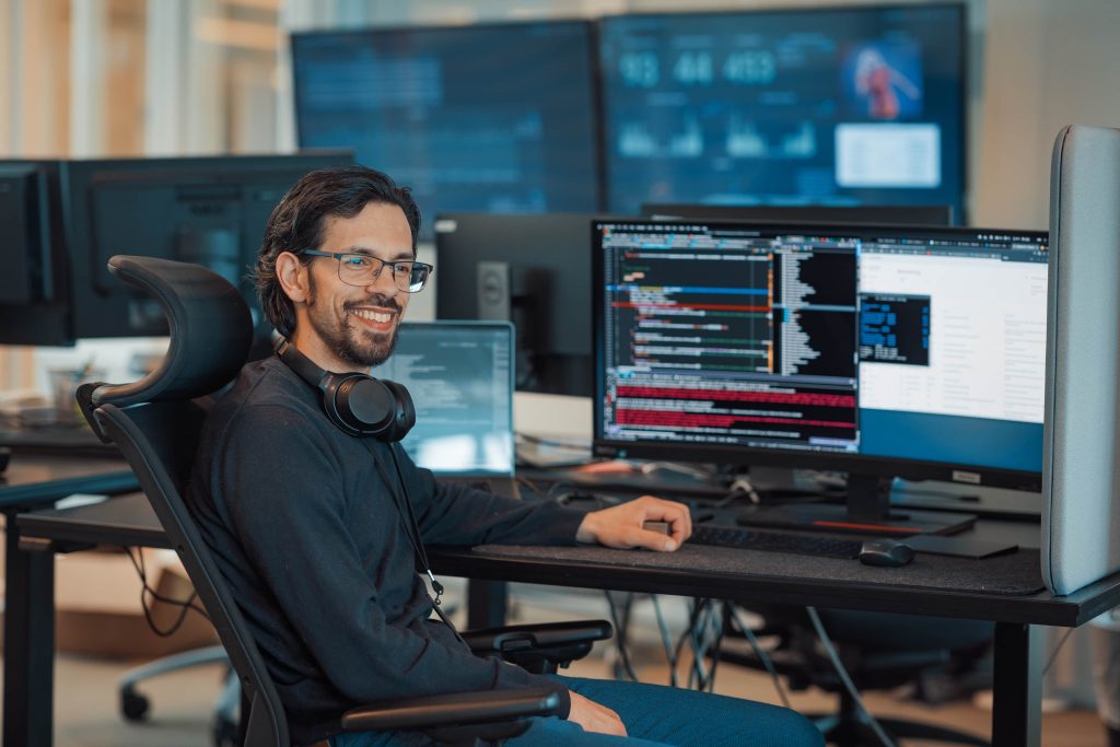 En mann med briller og hodetelefoner rundt halsen sitter ved et skrivebord med flere dataskjermer som viser kode i et kontormiljø.