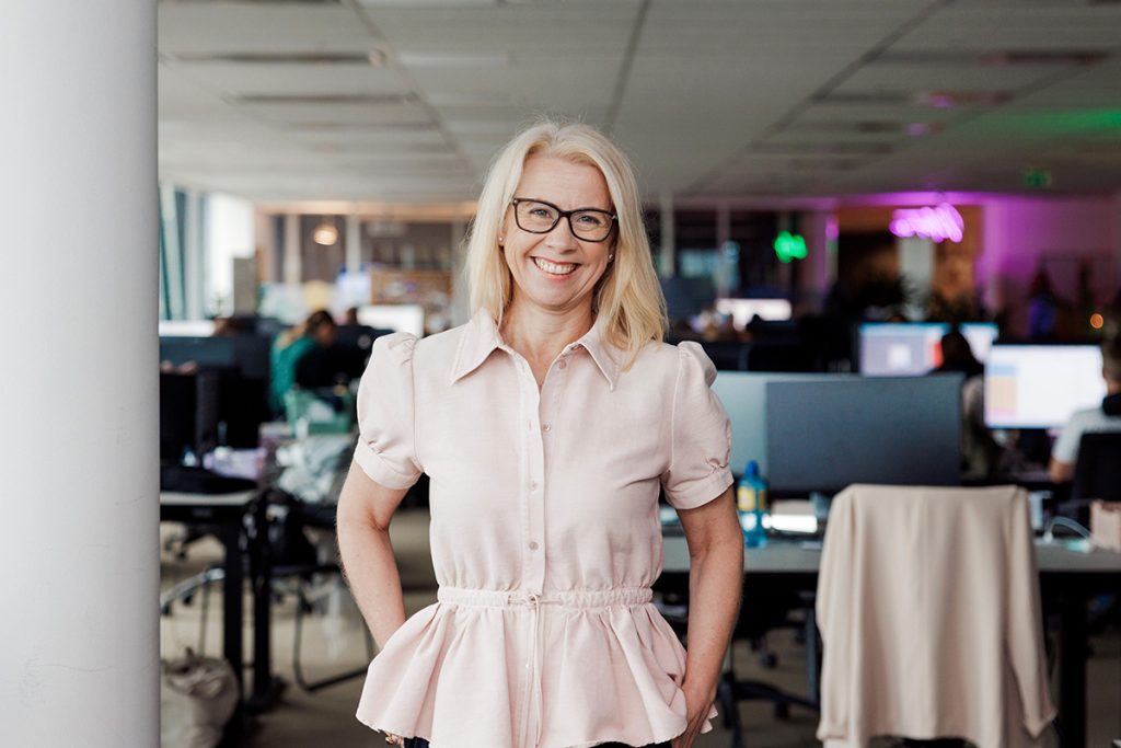 Lise Ekanger er direktør for strategiske kunderelasjoner i Tripletex. En kvinne med blondt hår og briller står smilende i et kontormiljø med pulter, skjermer og mennesker i bakgrunnen.