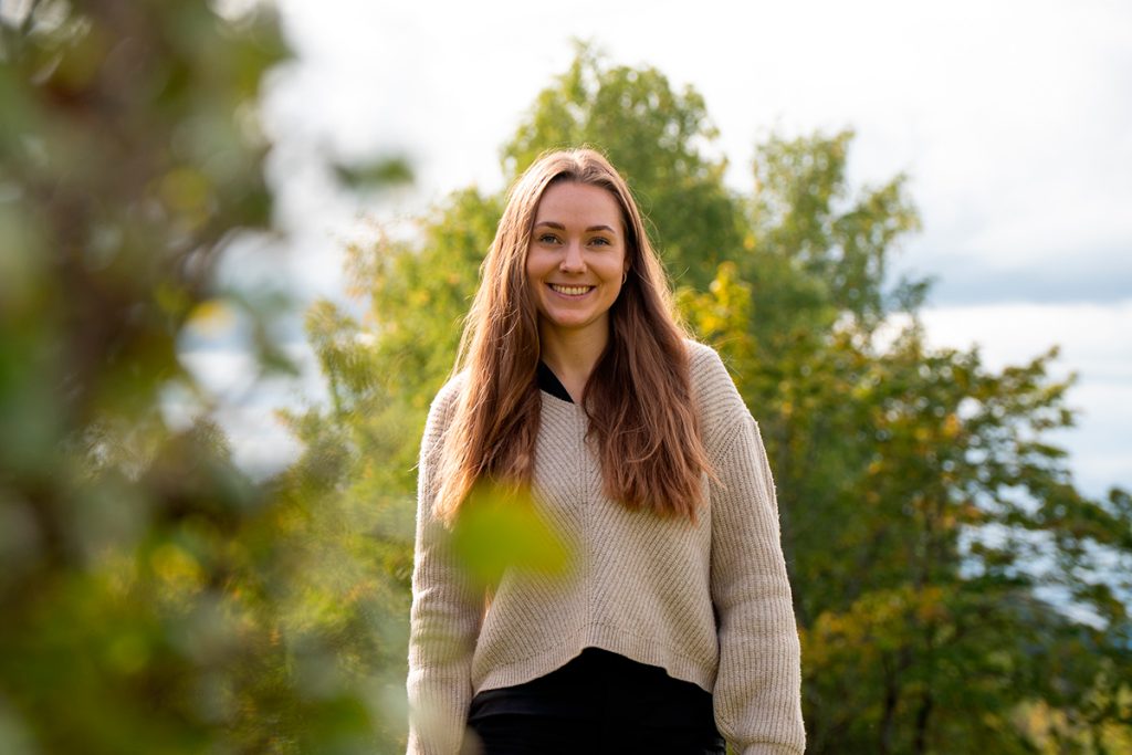 En kvinne med langt, brunt hår smiler utendørs, stående foran grønne trær og iført beige genser og svarte bukser. Jeanette Lystad snakker om bærekraft.