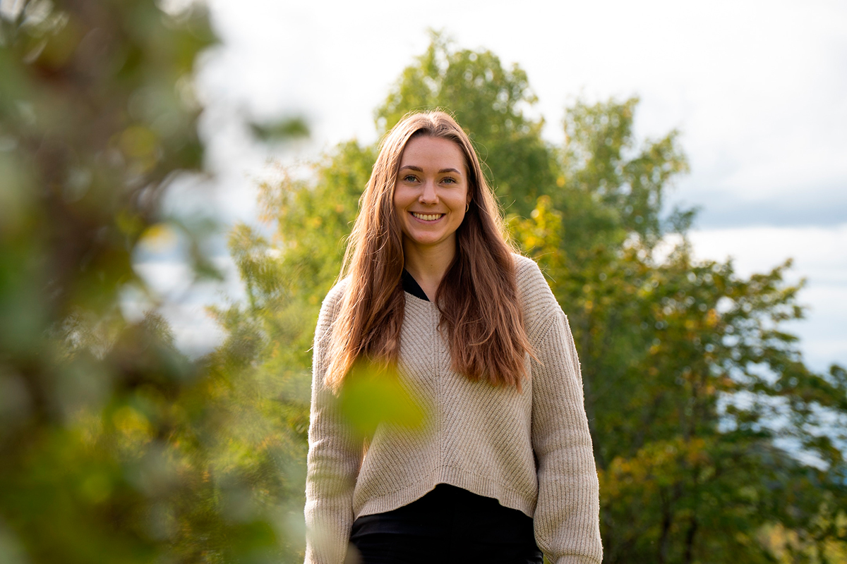 En kvinne med langt, brunt hår smiler utendørs, stående foran grønne trær og iført beige genser og svarte bukser. Jeanette Lystad snakker om bærekraft.