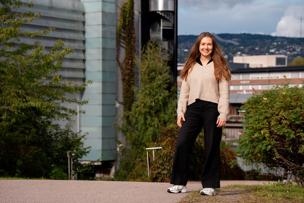 En kvinne med langt hår står utendørs på en asfaltert sti og smiler, med moderne bygninger og grøntområder i bakgrunnen. Jeanette Lystad snakker om bærekraft.