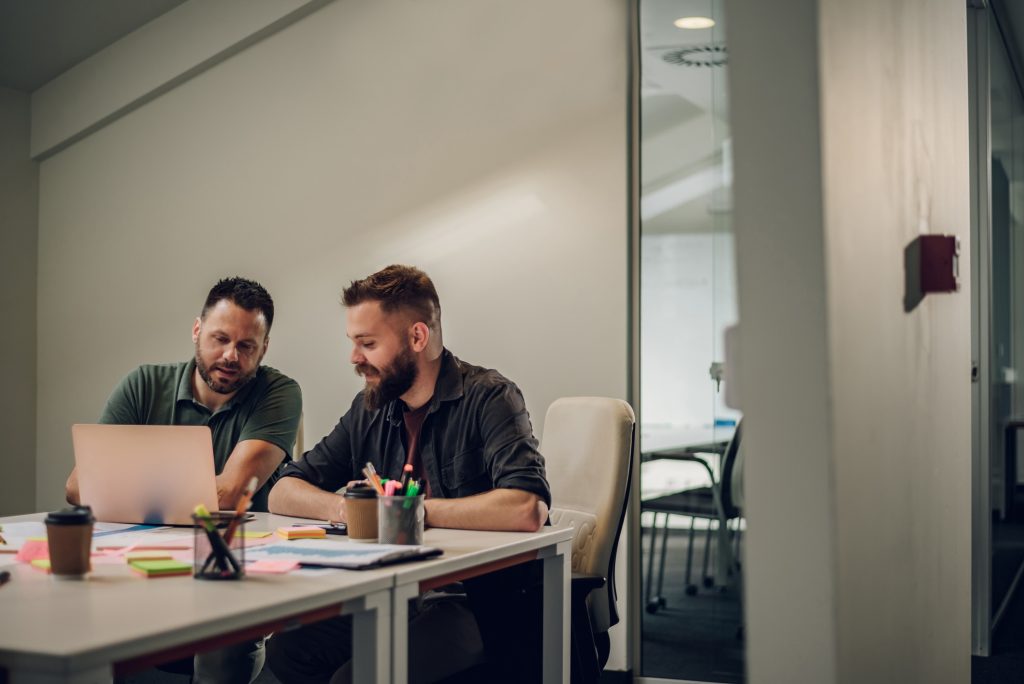 To menn sitter ved et skrivebord på et kontor og samarbeider mens de ser på en bærbar datamaskin og tar notater, med skrivesaker og kaffekopper på bordet.