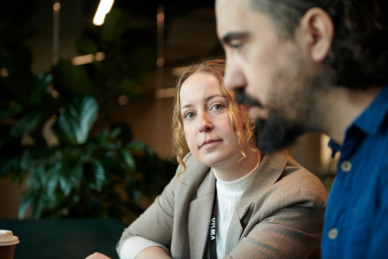 En kvinne i beige blazer ser inn i kameraet mens hun sitter ved siden av en mann i blå skjorte og muligens diskuterer løsninger for en kontantstrømskrise, med uskarpe grønne planter i bakgrunnen.