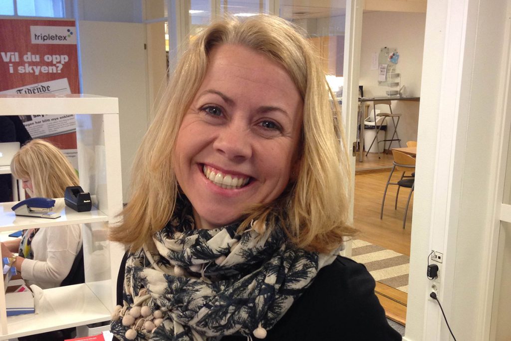 En kvinne med blondt hår smiler til kameraet inne på et kontor, iført mønstret skjerf og mørkeblå topp topp. Lise Ekanger er en av gründerne i Tripletex.